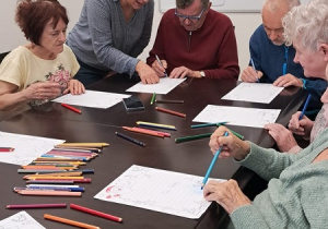 Grupa osób siedzi przy stole. Kolorują obrazki. Na stole leżą kredki.