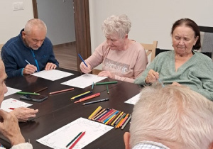 Grupa osób siedzi przy stole. Kolorują obrazki. Na stole leżą kredki.