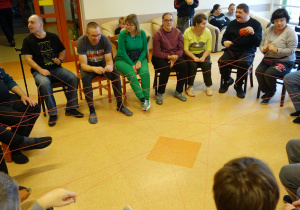 Grupa osób siedzących w okręgu przekazuje sobie kłębek wełny tworząc splątaną sieć.