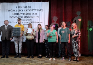 Grupa osób na scenie. Mężczyzna trzyma w rękach zapakowany prezent, jedna z kobiet dyplom, druga pamiątkową statuetkę.