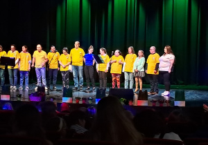 Grupa osób ubrana w żółte koszulki. Grupa stoi na scenie.