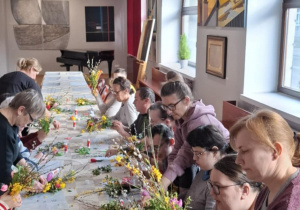Grupa osób siedzących przy stole. Wszyscy wykonują palmy wielkanocne.