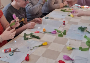 Grupa osób siedzących przy stole. Wszyscy wykonują palmy wielkanocne.