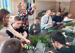 Grupa osób siedzących przy stole. Wszyscy wykonują palmy wielkanocne.