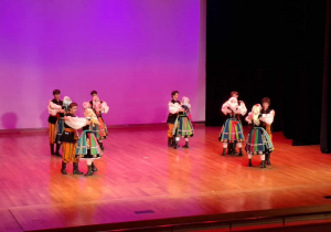 Grupa tancerzy ubrana w kolorowe stroje tańczy na scenie.