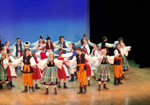 Grupa tancerzy ubrana w kolorowe stroje tańczy na scenie.