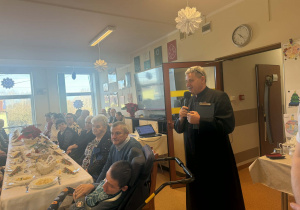 Grupa osób siedzących przy stole świątecznym. Obok do mikrofonu przemawia ksiądz ubrany w czarą sutannę.
