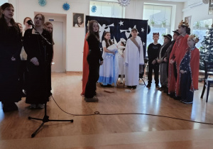 Występ teatralny. Grup osób na scenie, dwie dziewczynki ubrane na czarno śpiewają do mikrofonu. Na dalszym planie chłopiec i dziewczynka ubrani w białe szaty, dziewczynka trzyma w rękach lalkę symbolizującą małego Jezusa. Obok stoi grupa dzieci przebranych za pasterzy i i trzech królów.