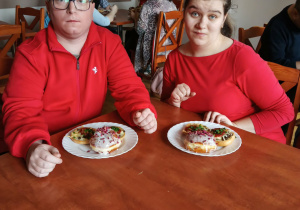 Kobieta i mężczyzna ubrani na czerwono siedzą przy stole, na którym znajdują się talerze z pączkami i zapiekankami.