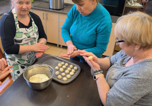 Trzy kobiety stoją przy stole, na którym stoi metalowa misa i taca kulkami z ciasta. Kobiety formują kulki z ciasta.