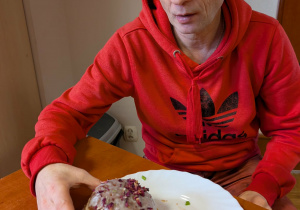 Mężczyzna w czerwonej bluzce siedzi przy stole, na którym znajduje się biały talerz z pączkiem.