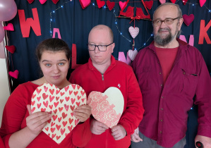 Kobieta i dwóch mężczyzn. Kobieta i jeden z mężczyzn ubrani są w w kolorach czerwieni. Kobieta i mężczyzna trzymają w rękach walentynkowe serca. Wszyscy znajdują się na tle granatowego parawanu, który jest ozdobiony walentynkowymi sercami.