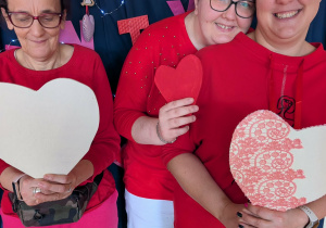 Trzy kobiety ubrane w czerwone bluzki trzymają w rękach walentynkowe serca.