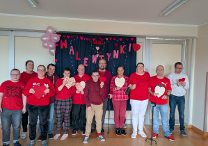 Grupa osób ubranych w czerwone bluzki symbolizujące święto zakochanych. Większość osób trzyma w rękach walentynkowe serca. Grupa stoi na tle granatowego parawanu z napisem walentynki, który jest przyozdobiony czerwonymi sercami i balonami.