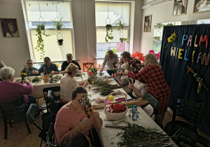 Grupa osób wykonuje palmy wielkanocne. Na stole leżą materiały wykorzystywane do zrobienia palmy wielkanocnej.