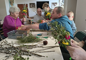 Kilka osób tworzy palmy wielkanocne. Osoby siedzą przy stole, na który znajdują się materiały wykorzystywane do zrobienia palmy wielkanocnej.