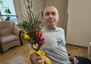 Mężczyzna w szarej koszulce siedzi na krześle, w ręku trzyma wykonana przez siebie palmę wielkanocną.