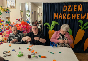 Grupa trzech osób siedzących przy stole, na którym znajduja cię materiały do wykonania breloczka w kształcie marchewki.