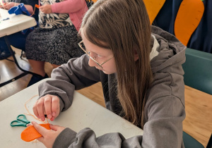 Dziewczynka siedzi przy stole, w rękach trzyma igłę oraz materiał, z którego wykonuje marchewkowy breloczek.