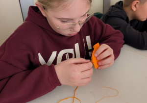 Dziewczynka siedzi przy stole, w rękach trzyma igłę oraz materiał, z którego wykonuje marchewkowy breloczek.
