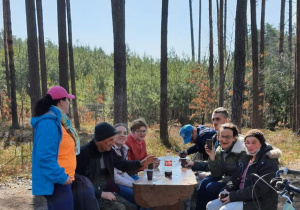 kobiety i męzczyżni siedzą przy stole na leśnej polanie