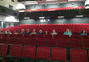 Trzynaście kobiet i mężczyzn siedzących w jednym rzędzie w sali kinowej na czerwonych fotelach