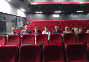 Siedem osób kobiet i mężczyzn siedzących na czesrwonych fotelach w kinie w jednym rzędzie