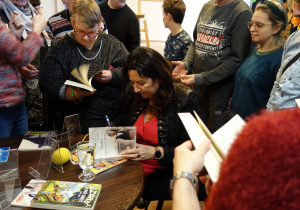 Grupa osób stojących wokół stolika, przy którym siedzi kobieta podpisująca książkę