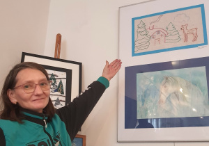 Kobieta w okularach unosząca dłoń wskazującą na ścianę, gdzie wisi obraz