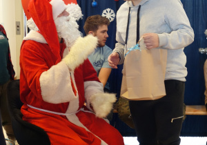 Św. Mikołaj ubrany w czerwony strój i czapkę ofiarowuje mężczyźnie prezent w szarej torbie