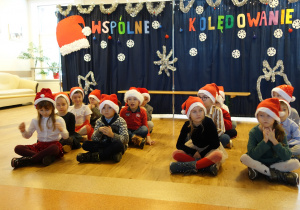 Grupa dzieci w biało-czerwonych czapkach z białymi pomponami, siedzi po turecku na podłodze. Za nimi granatowy parawan przystrojony świątecznie