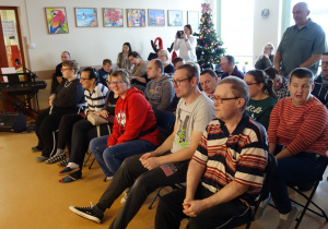 Grupa osób siedzących na krzesłach ogląda występ świąteczny, w tle bożonarodzeniowa choinka