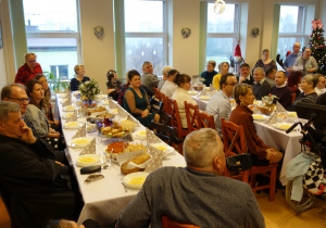 Grupa ludzi siedzących przy świątecznych stołach, Na stołach białe obrusy i potrawy bożonarodzeniowe