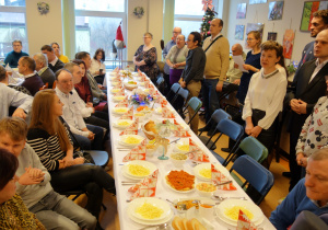 Przy zastawionym świątecznymi potrawami stole siedzi grupa osób. Przed stołem stoi grupa osób biorąca udział w występie artystycznym