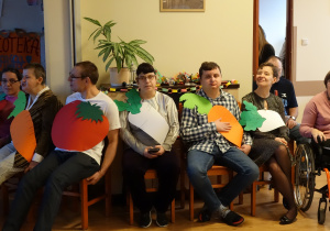 Grupa osób siedzi na krzesłach. Każda osoba trzyma warzywo lub owoc wykonane z kolorowych kartonów