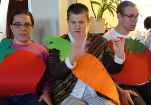 Grupa trzech osób siedzi na krzesłach, każda trzyma warzywa wykonane z kolorowych kartonów