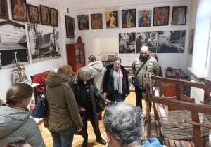 Grupa osób zwiedza muzeum. W pomieszczeniu znajdują się eksponaty a na ścianach wiszą obrazy.