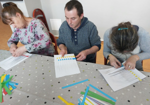 Dwie kobiety i mężczyzna siedzą przy stole wykonując pracę plastyczną z użyciem białej kartki i kolorowych pasków papieru