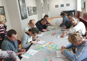 Grupa osób siedzących przy stole wykonuje pracę plastyczną z kolorowych pasków na białej kartce papieru