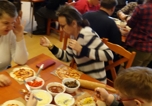 Duża grupa kobiet i mężczyzn przy stołach dekorują pizzę