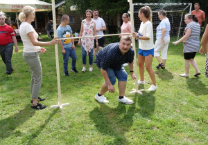 Zabawa w przechodzeniu pod drewnianą poprzeczką, która jest umieszczana na różnych poziomach. Dwie kobiety podtrzymują drewnianą poprzeczkę, pod którą przechodzi schylony mężczyzna.